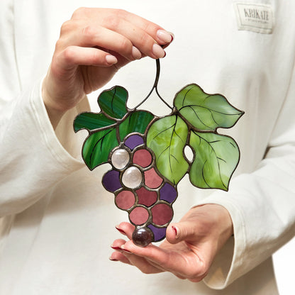 A person holding a handcrafted stained glass suncatcher shaped like a grape branch with purple grapes and green leaves.