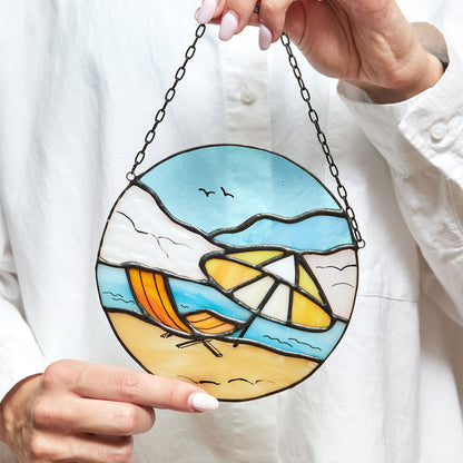 A hand holding a round stained glass suncatcher with a beach and ocean landscape design, featuring blue sky, white clouds, yellow sun, and a multicolored sea.