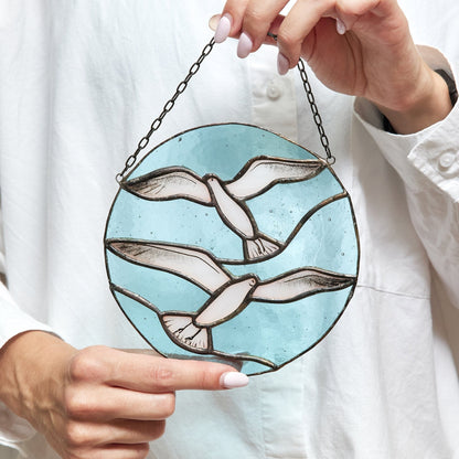 A hand holding a round stained glass suncatcher with a seagull design, predominantly in shades of blue and white, with the piece yet to be hung.