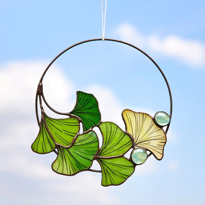 A circular stained glass window hanging featuring green and yellow Ginkgo Biloba leaf patterns with small blue accents, suspended against a blue sky background.