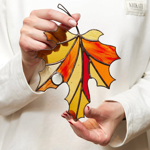 A person holding a handcrafted stained glass suncatcher shaped like a maple leaf with autumn colors of red, orange, and yellow.