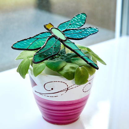 A handcrafted stained glass suncatcher shaped like a dragonfly, displayed on top of a potted plant.