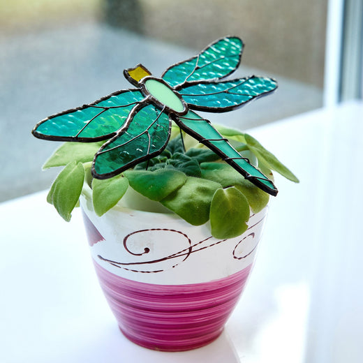 A handcrafted stained glass suncatcher shaped like a dragonfly, displayed on top of a potted plant.