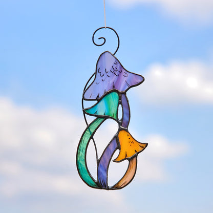 A colorful stained glass suncatcher in the shape of a mushroom with a bird perched on it, hanging against a blue sky with clouds.