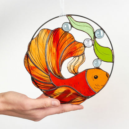 Handmade stained glass suncatcher in the shape of a goldfish with bright orange and green colors, held in a person's hand.