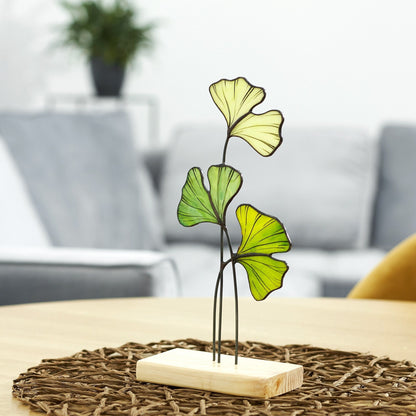 A handcrafted stained glass Ginkgo Leaf table stand decoration with a metal stand, placed on a wooden platform, set against a cozy home background.