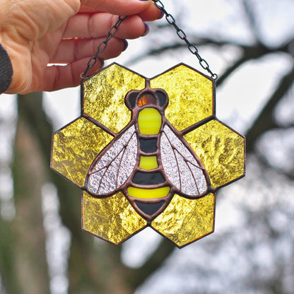 A handmade stained glass suncatcher featuring a yellow honeycomb pattern with a central bee design, held against an outdoor backdrop.