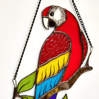 A handmade stained glass suncatcher in the shape of a colorful parrot, with red, blue, and green glass, perched on a branch.