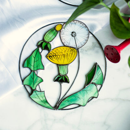 Handmade stained glass suncatcher in the form of a dandelion flower with a yellow petal and seed head, green leaves, and a circular frame.