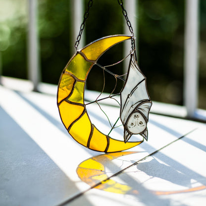 A stained glass decoration featuring a yellow crescent moon with a black bat in the center, hanging by a chain.