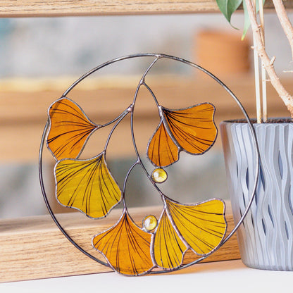 A handcrafted stained glass suncatcher in the shape of Ginkgo Biloba autumn leaves, with yellow and orange leaves and a copper foil structure, placed on a wooden surface.