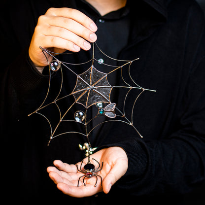 A person holding a handcrafted stained glass decoration featuring an intricate spider web with a spider and marbled glass water droplets.